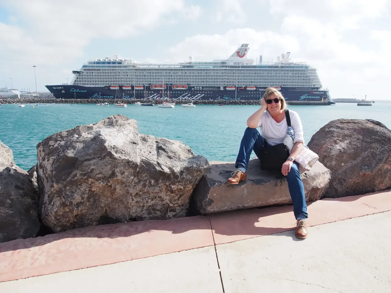 Eine Frau sitzt am Hafen auf einem Felsen, dahinter liegt ein großes Kreuzfahrtschiff